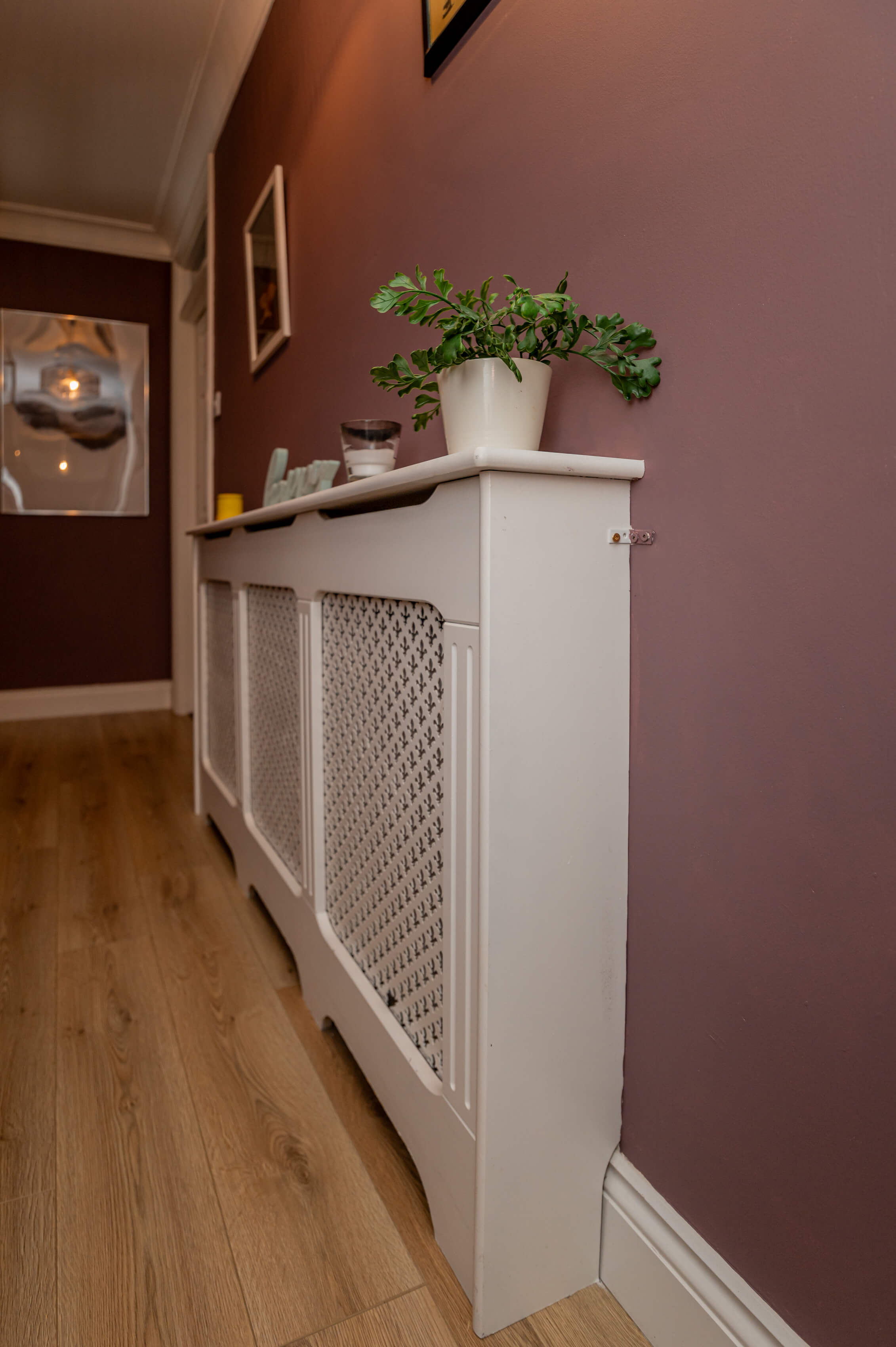 a hallway with a white radiator cover, and dusky purple walls. Photographs and ornaments on the radiator cover and pictures on the walls.